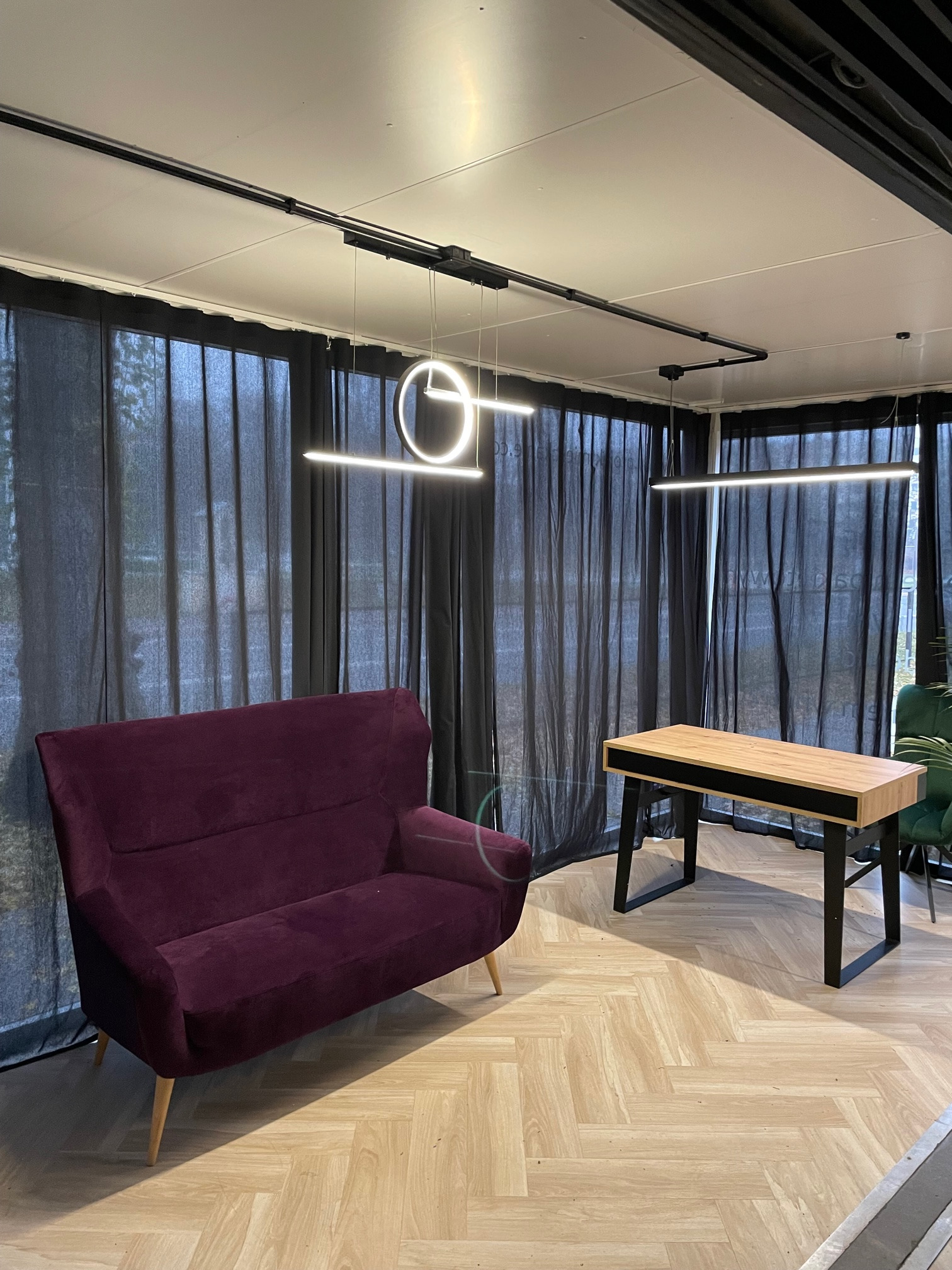 Nowoczesne wnętrze biurowe z bordową sofą, drewnianym biurkiem z czarnymi nogami, panelami podłogowymi w jodełkę i minimalistycznymi lampami wiszącymi na tle zasłon.