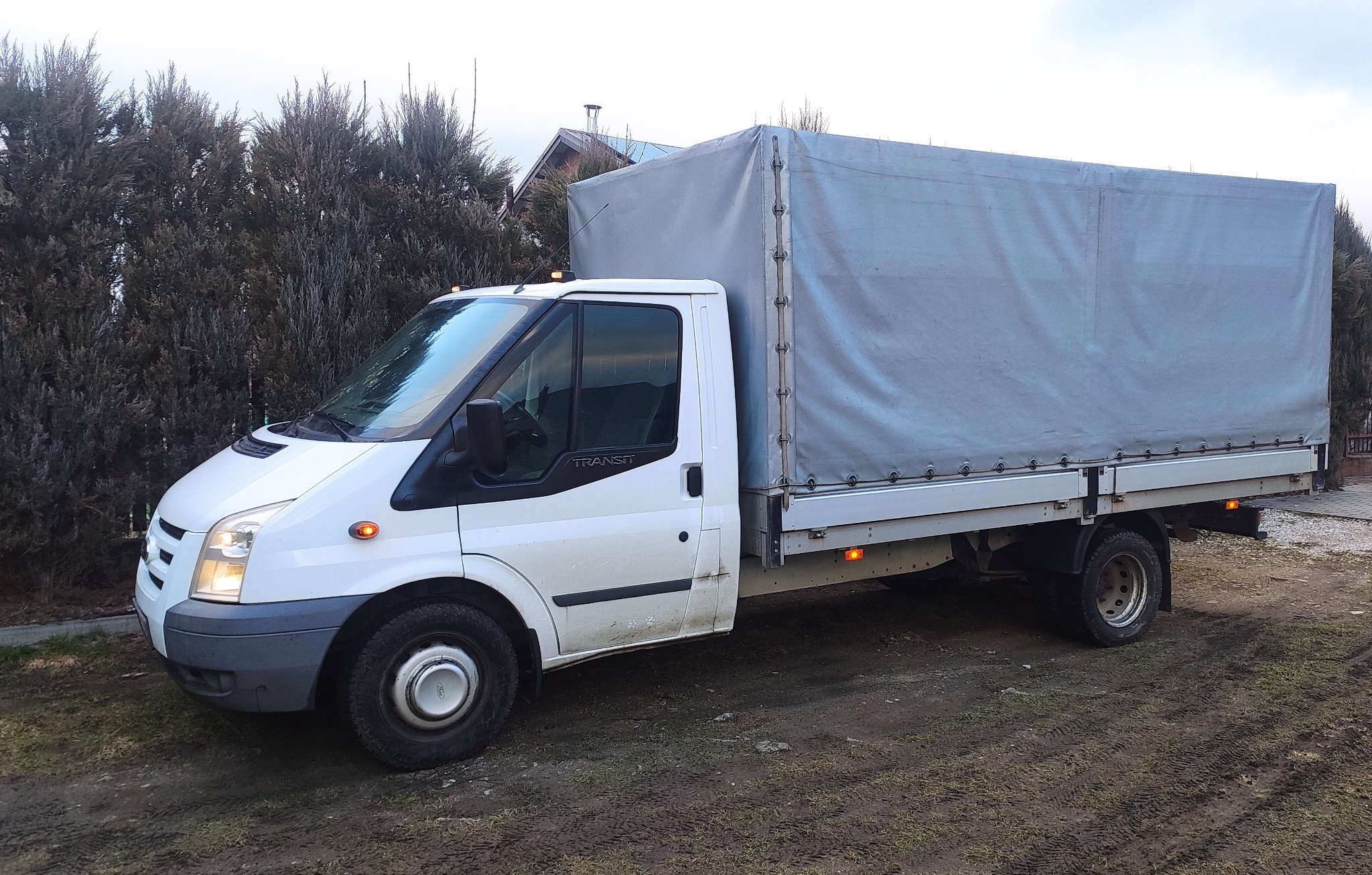 Biały samochód dostawczy z plandeką na błotnistym podłożu, widoczne ślady użytkowania na karoserii.
