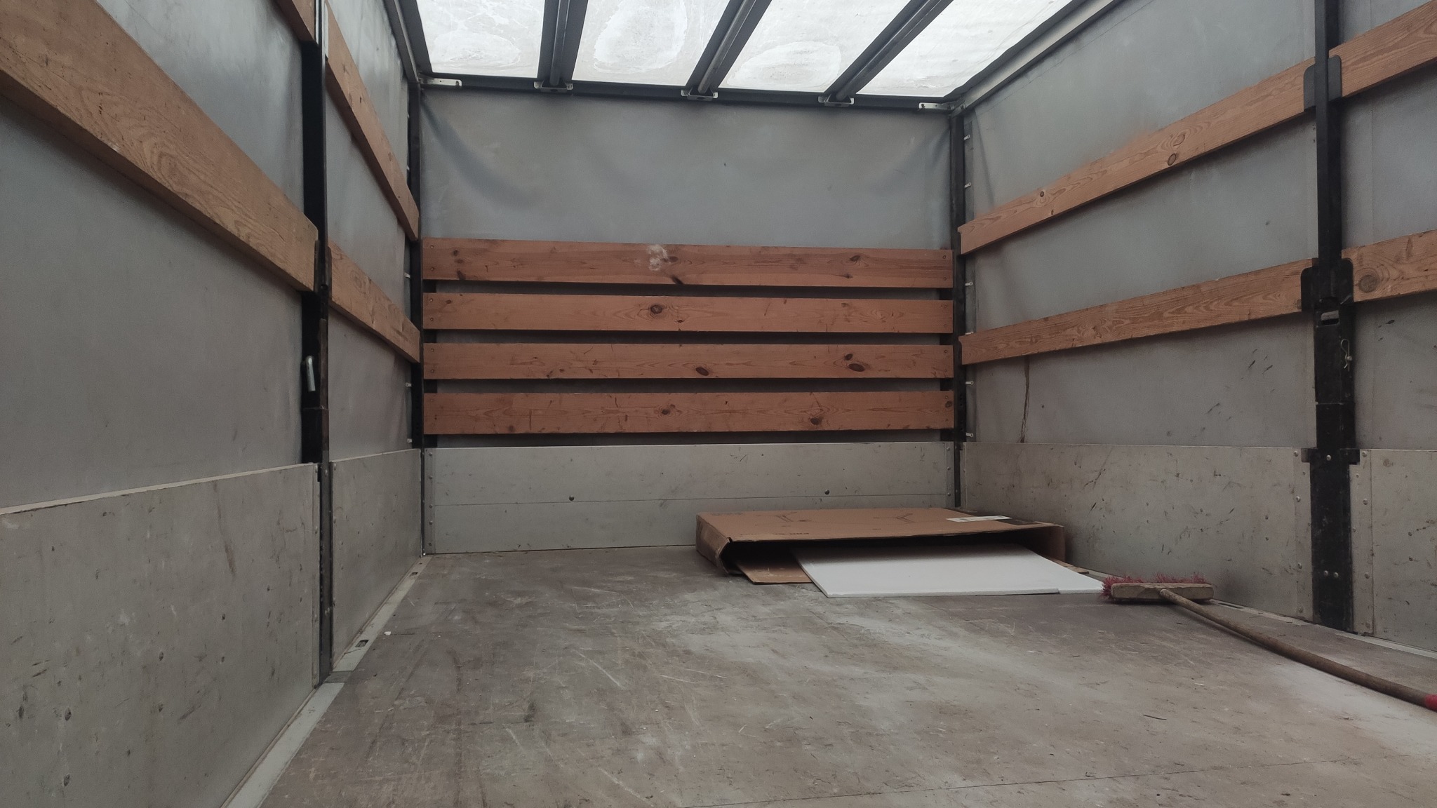 Wnętrze paki pojazdu dostawczego z drewnianymi listwami zabezpieczającymi ładunek, kartonem i zmiotką na podłodze.