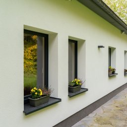 Robstan Producent Okien - Elewacja domu z trzema oknami z czarnymi ramami, na parapetach doniczki z żółtymi kwiatami i wrzosem, widok z boku na kamienny chodnik i fragment ogrodu.