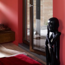 Robstan Producent Okien - Nowoczesne okno z ciemnobrązową ramą w pokoju z czerwoną ścianą, obok drewniana rzeźba afrykańska.