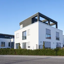 Nowoczesny, trzypiętrowy dom z białymi ścianami i szarym dachem, z szarymi roletami zewnętrznymi opuszczonymi w oknach, otoczony niskim żywopłotem.