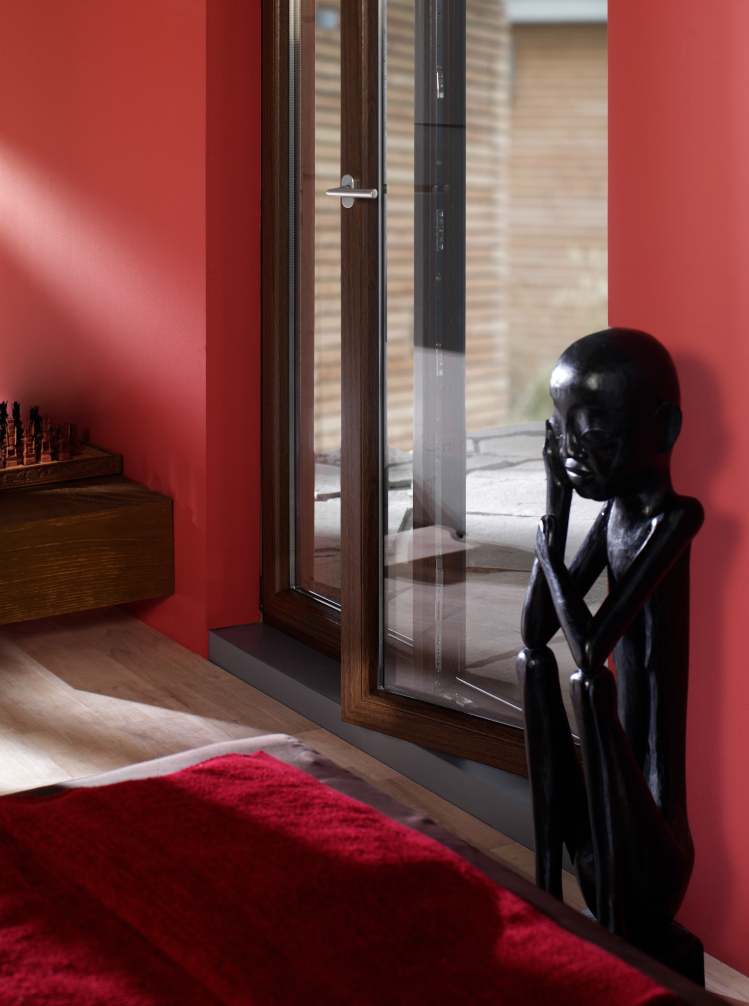 Nowoczesne okno z ciemnobrązową ramą w pokoju z czerwoną ścianą, obok drewniana rzeźba afrykańska.