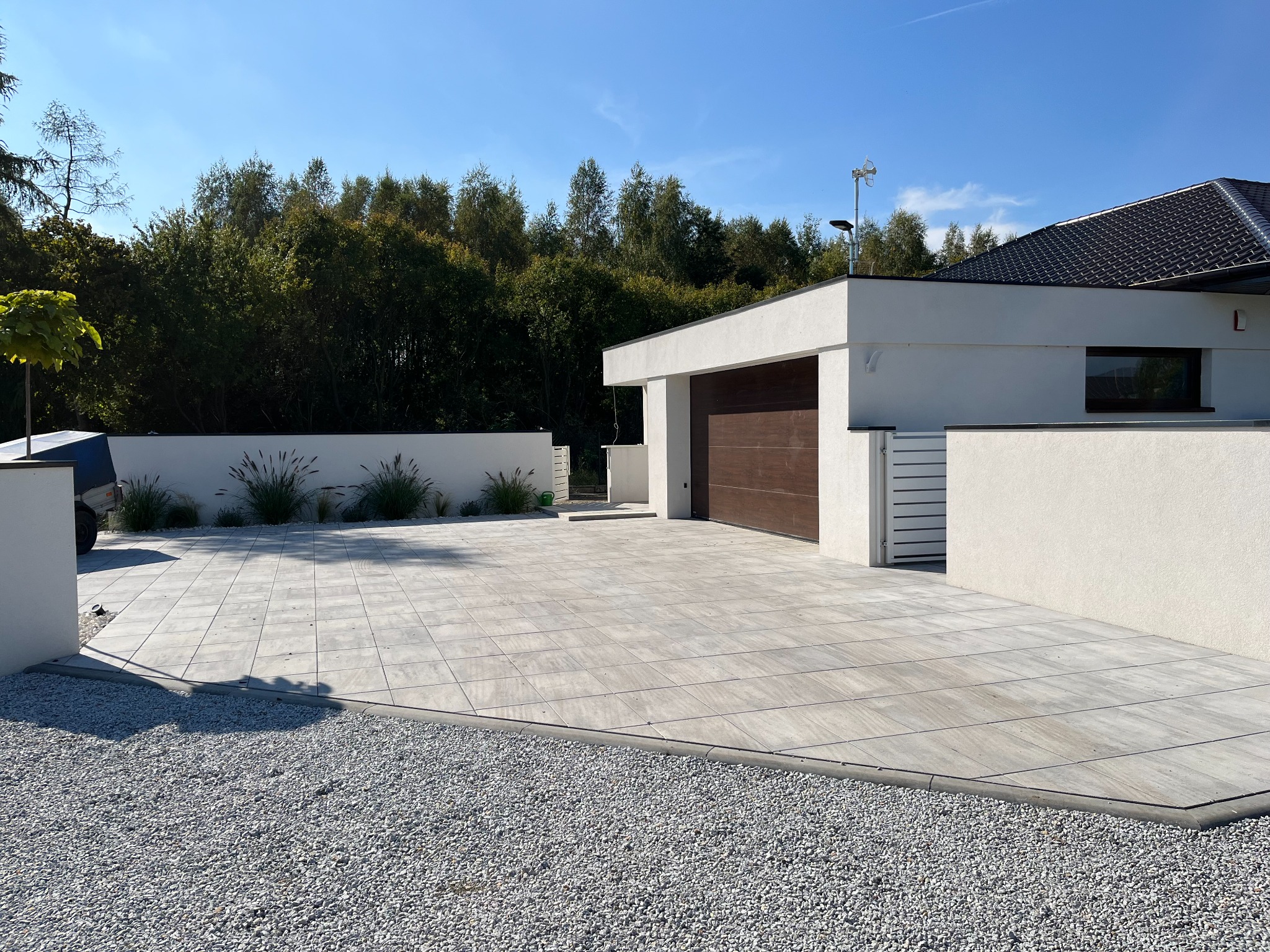 Nowoczesny podjazd z jasnoszarej kostki brukowej ułożonej w regularny wzór, obrzeża wykończone szarym krawężnikiem, a obok biały żwir i minimalistyczny dom z drewnianą bramą garażową.