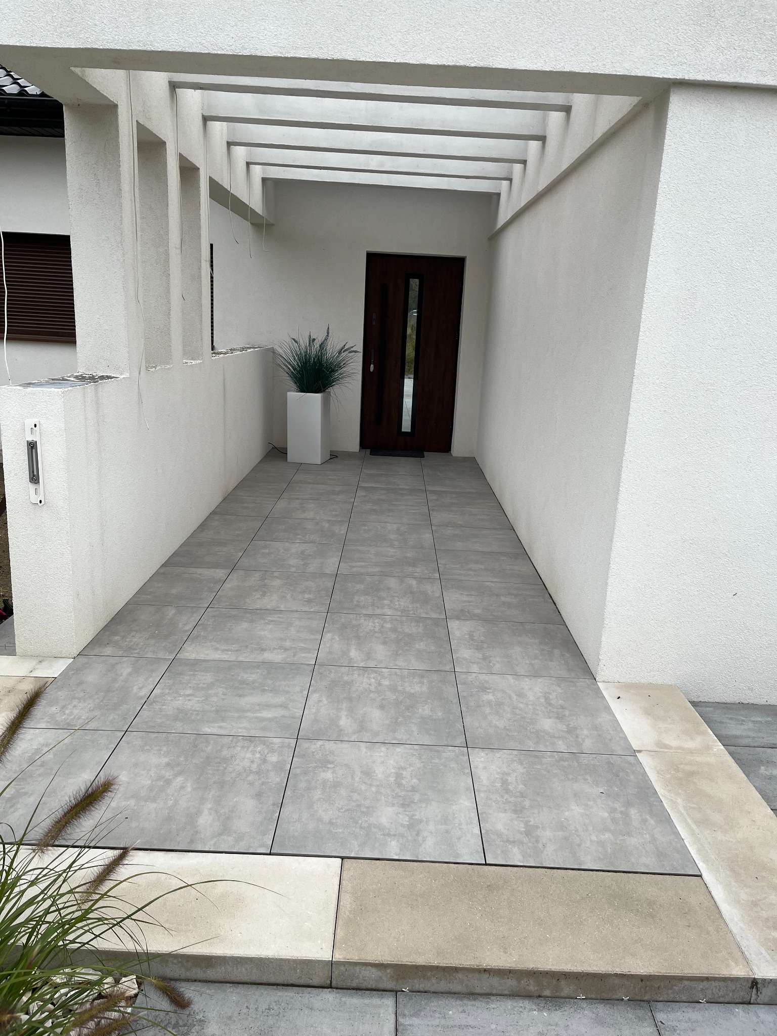 Nowoczesne wejście do domu z szarych, kwadratowych płyt betonowych, minimalistyczna aranżacja z trawą ozdobną w białej donicy i białymi ścianami.