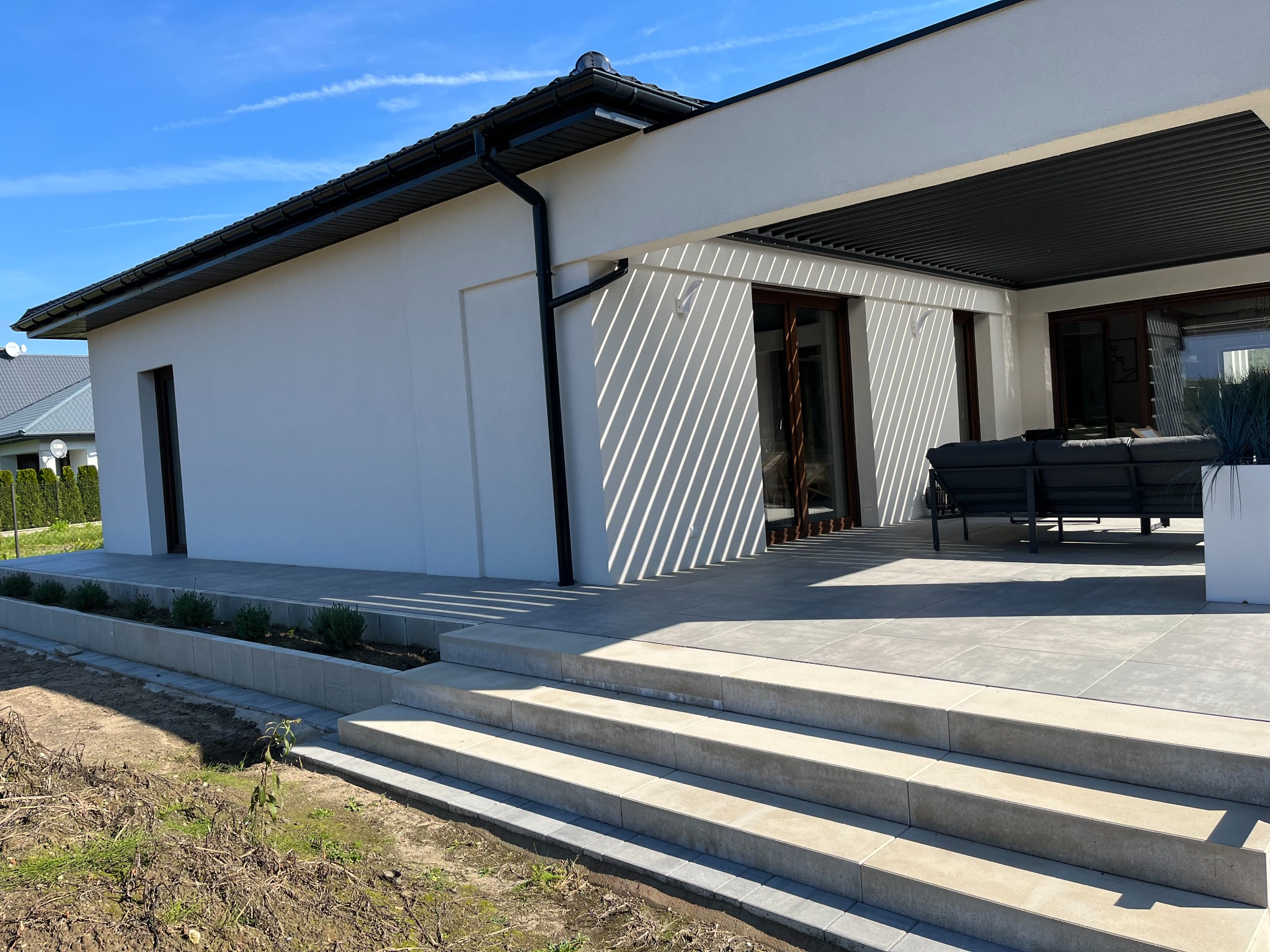 Nowoczesny taras z jasnoszarych płyt, schody z kostki w odcieniach beżu, minimalistyczna architektura domu w tle, rabata z zielenią wzdłuż ściany.