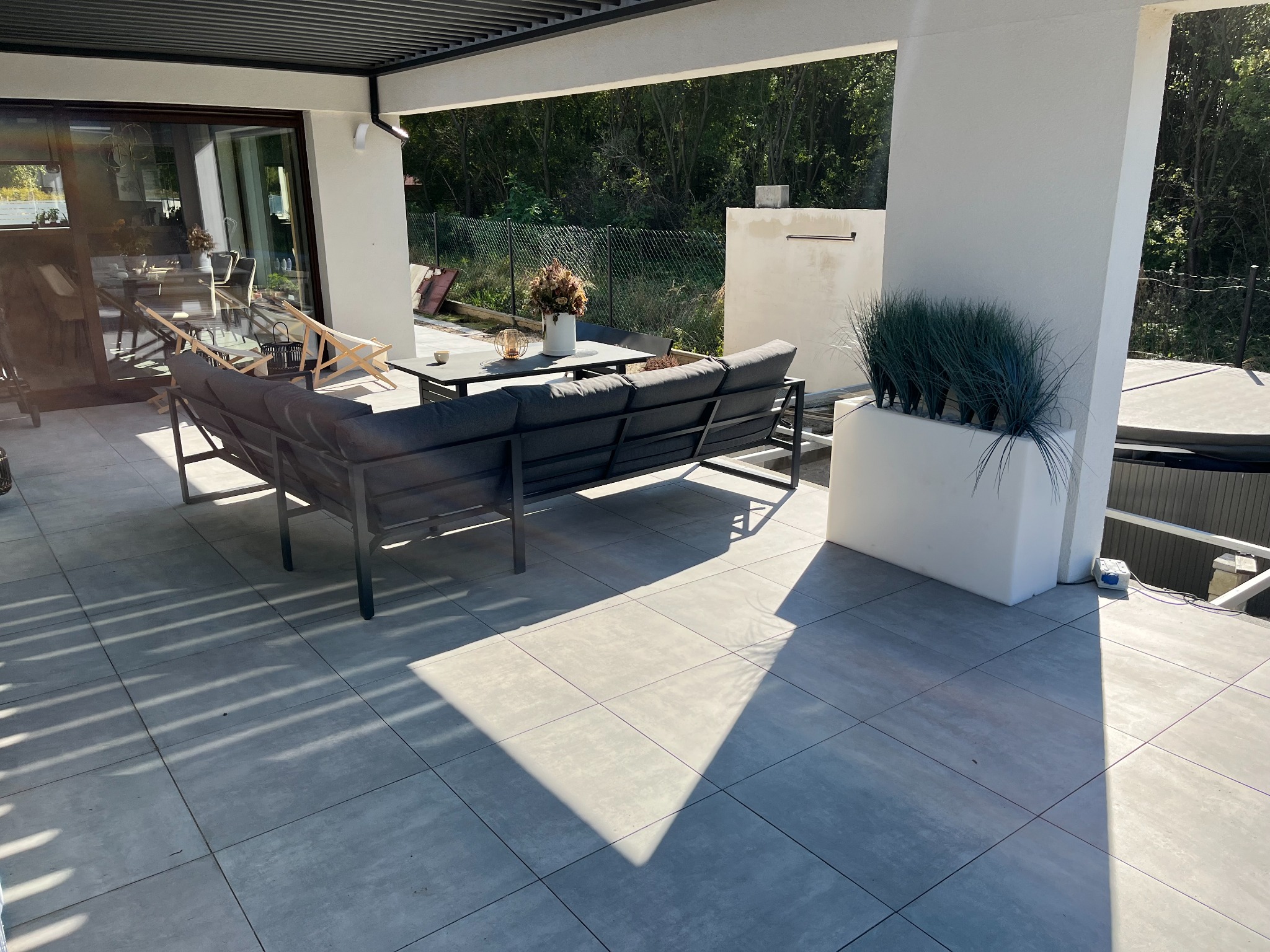 Jasnoszare płytki tarasowe ułożone na zewnątrz, w cieniu pergoli, z widocznym zestawem mebli ogrodowych i dekoracyjną roślinnością w donicy.