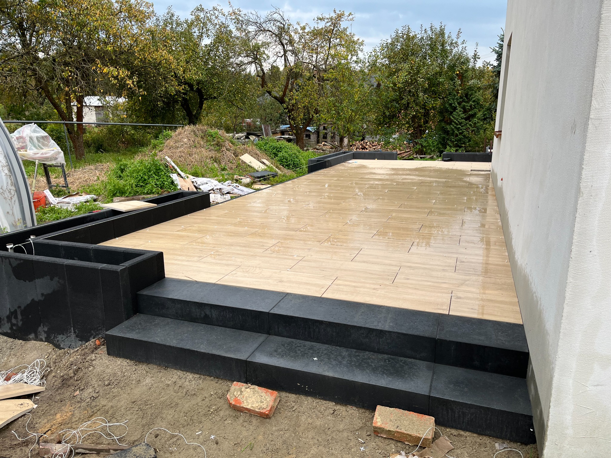 Ułożona, jasna kostka brukowa imitująca drewno na tarasie z czarnymi obrzeżami i schodami, widoczne ślady wilgoci po deszczu, w tle ogród z drzewami.