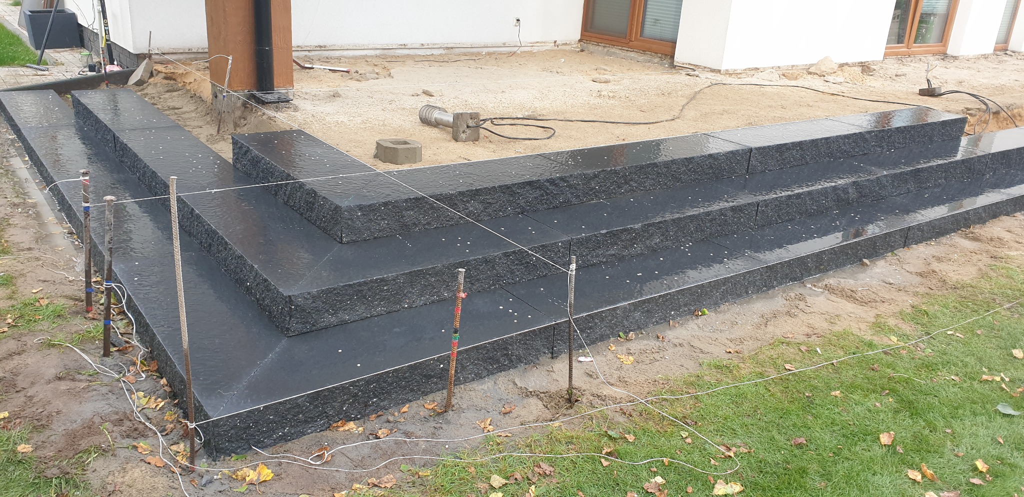 Nowo ułożone, ciemne, granitowe schody zewnętrzne z kostki brukowej, widoczne szpilki i linki wyznaczające poziom, w tle piasek i trawa.