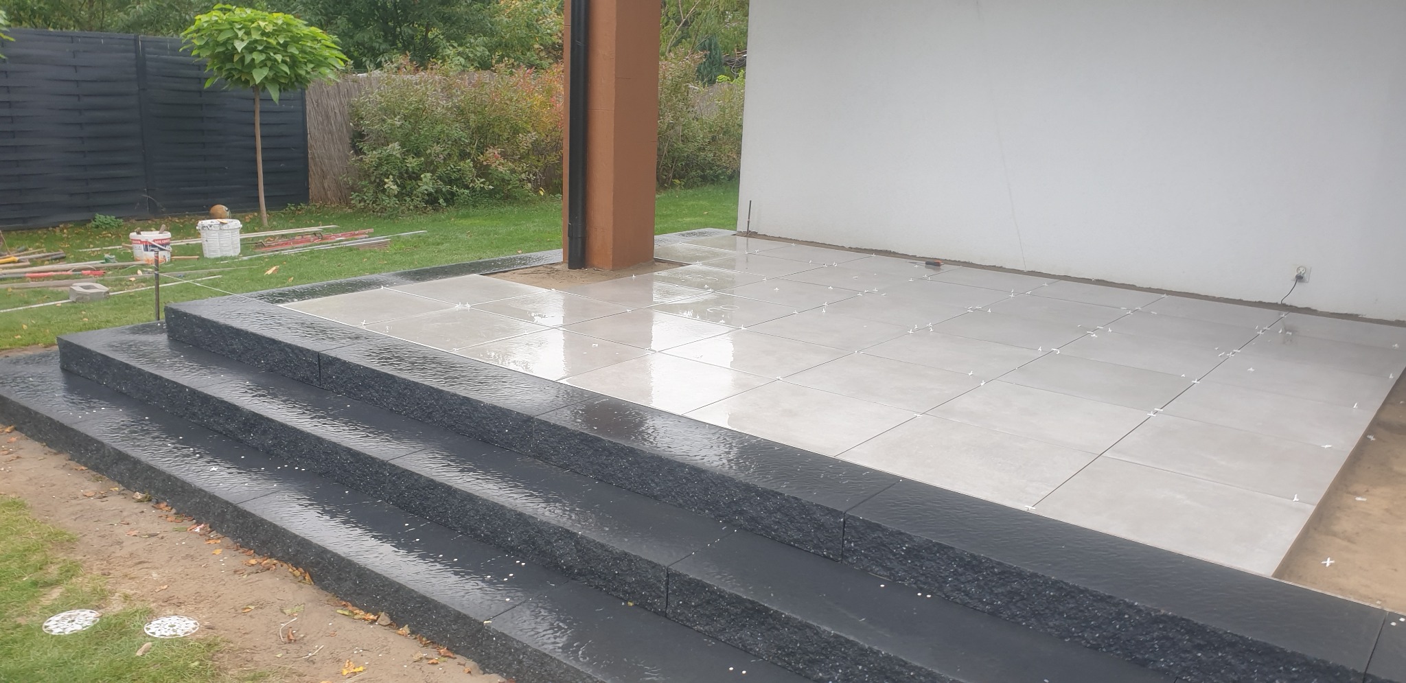 Montaż jasnych płytek na tarasie z ciemnymi schodami, widoczne krzyżyki dystansowe, słup podtrzymujący zadaszenie.