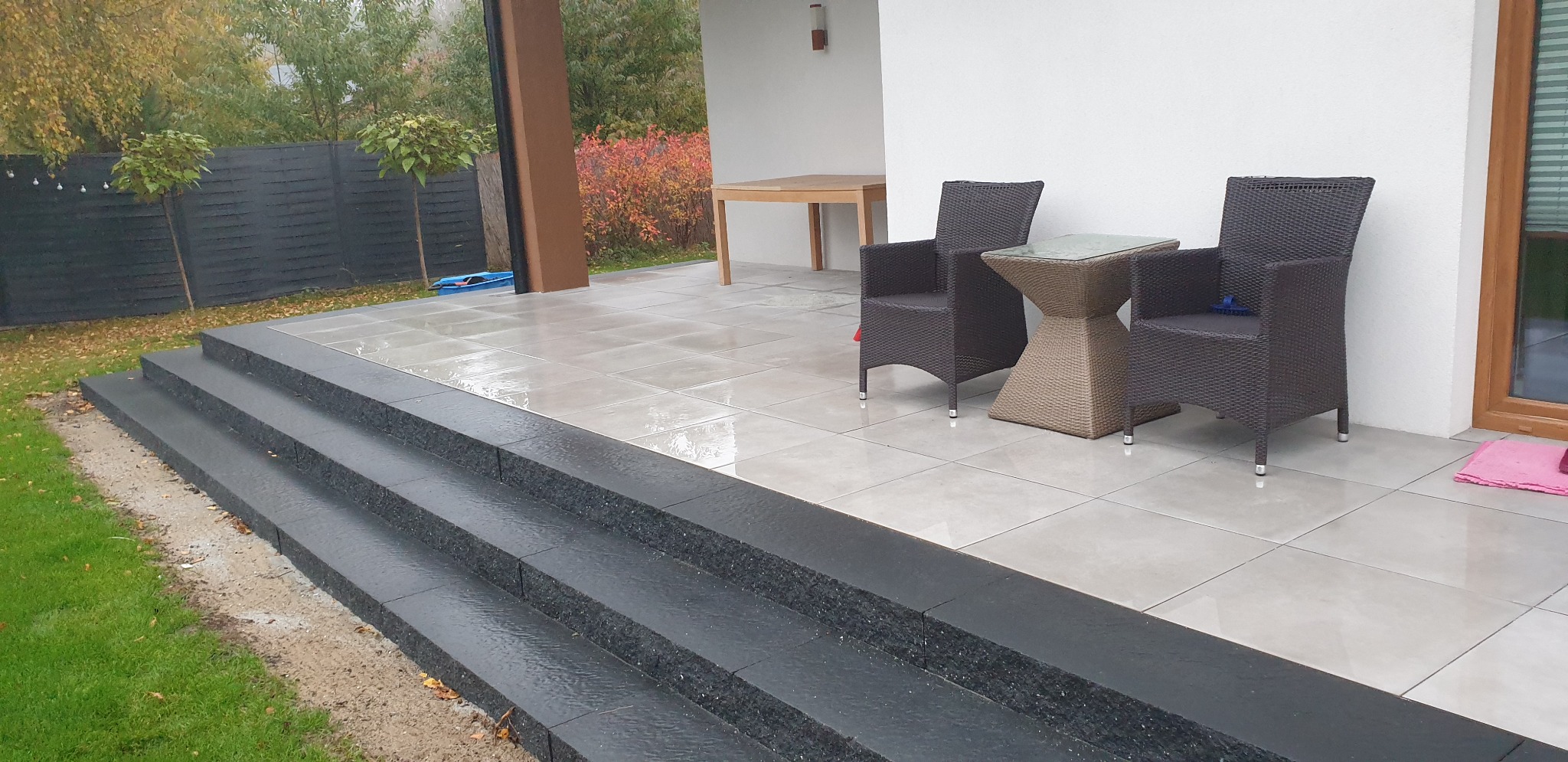 Wykończony taras z szarych płyt z czarnymi schodami, meble ogrodowe z rattanu, jesienny krajobraz w tle.