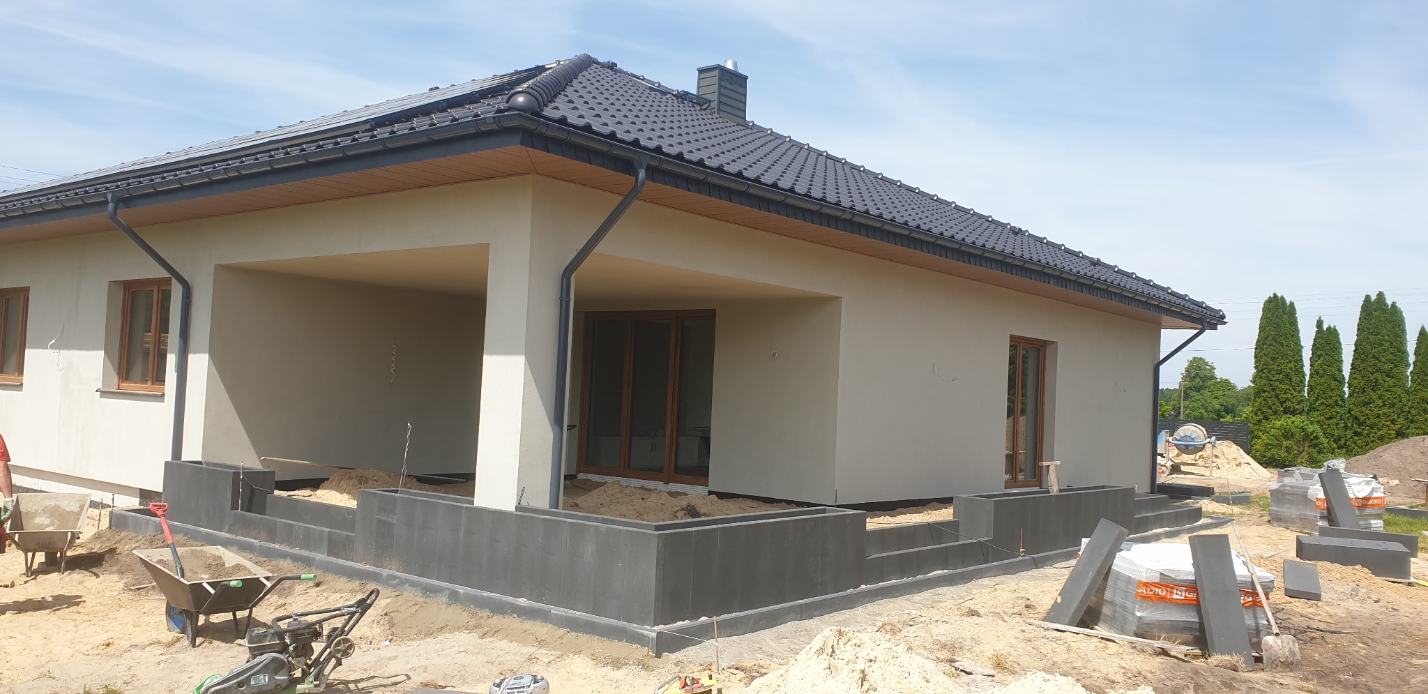 Nowoczesny dom jednorodzinny w trakcie budowy, widoczne fundamenty z szarej kostki, przygotowane podłoże i materiały budowlane, w tle wysokie tuje.