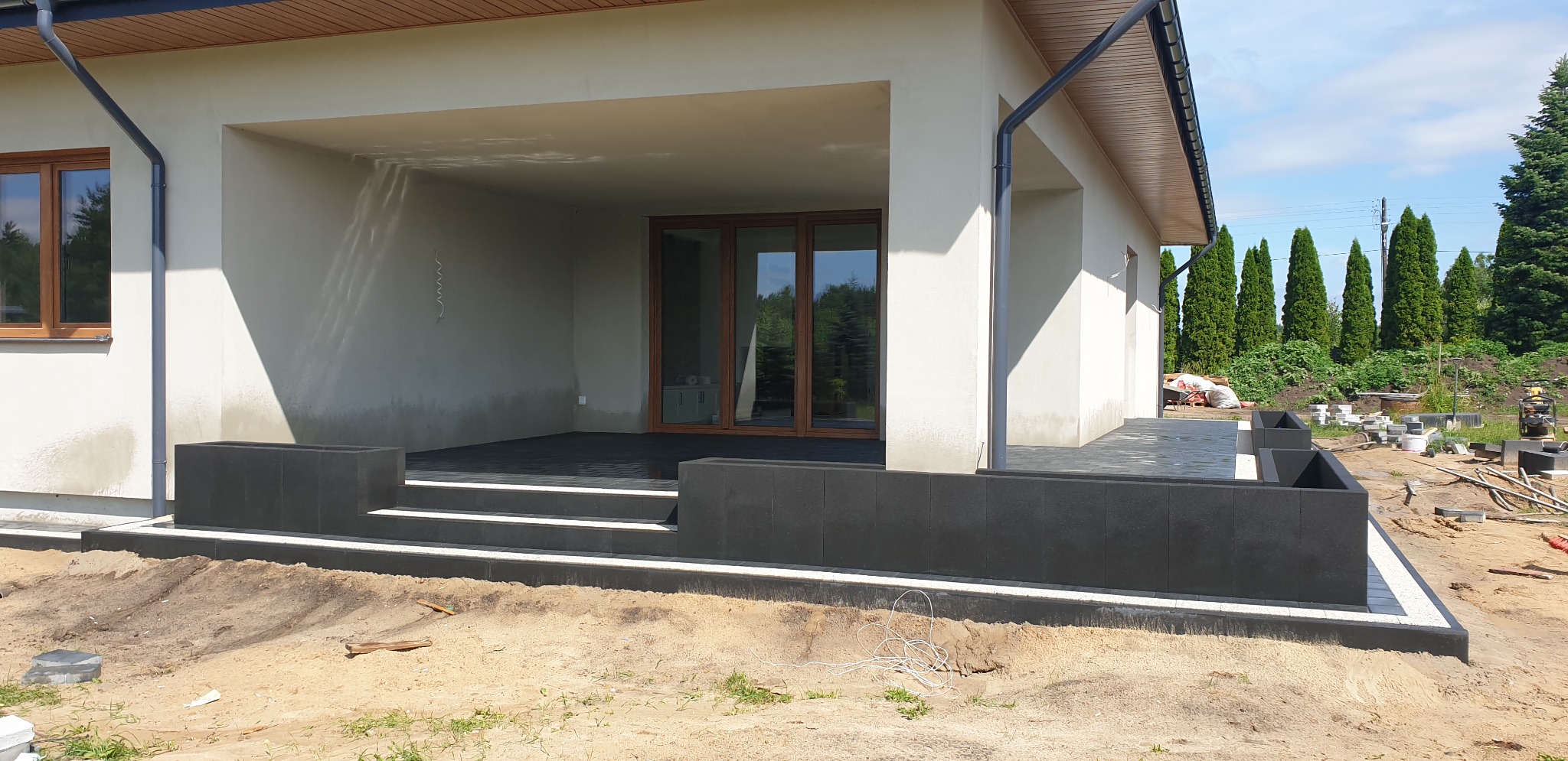 Nowoczesny taras z ciemnej kostki brukowej przed domem, widoczne schody z jasnymi obrzeżami i donice z tej samej kostki, w tle wysokie tuje.