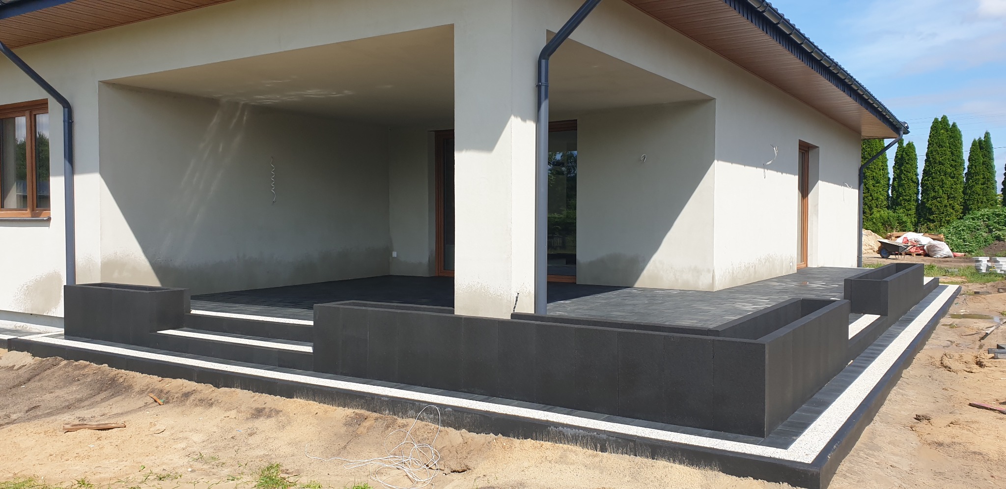 Nowoczesny taras z ciemnoszarą kostką brukową, obrzeżami z białego grysu i donicami, widok na dom z zewnątrz, w tle tuje.