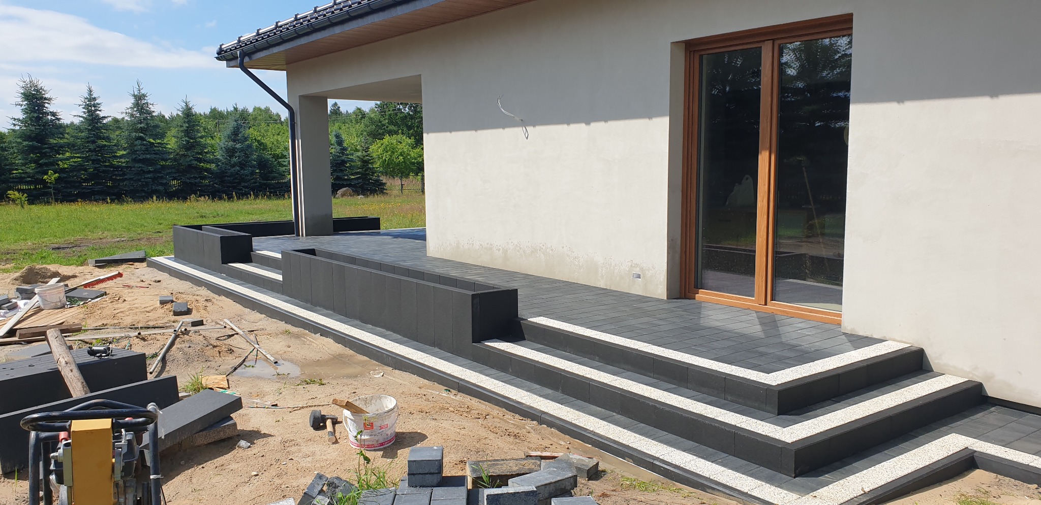 Częściowo ułożona, ciemnoszara kostka brukowa na tarasie z donicami, widoczny zagęszczarka, wiadro i narzędzia, w tle dom jednorodzinny.