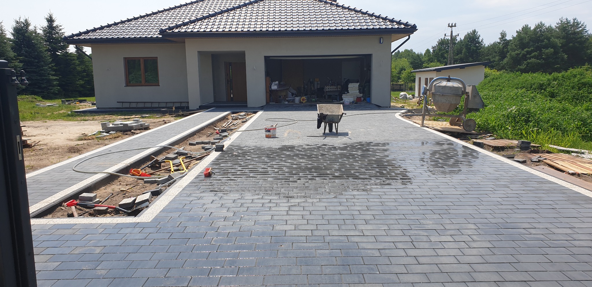 Częściowo ułożona, mokra kostka brukowa w kolorze grafitowym tworząca podjazd do nowoczesnego domu z garażem; widoczne narzędzia brukarskie, taczka i betoniarka.