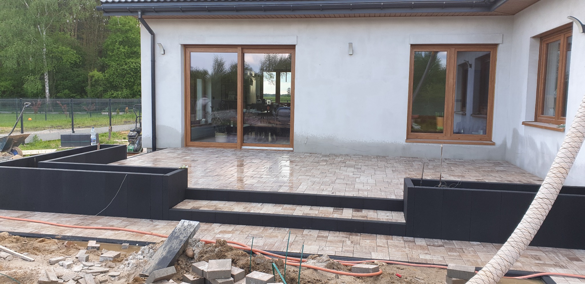 Tarasy z beżowej kostki brukowej ułożone przed domem z brązowymi oknami, widoczne czarne donice i elementy w trakcie budowy.