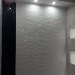 Wykończona ściana łazienki z białymi, trójwymiarowymi płytkami o falistym wzorze, sąsiadująca z czarną mozaiką i szarymi płytkami z wnękami podświetlanymi punktowym oświetleniem.