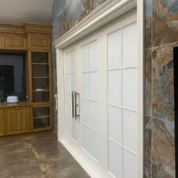 Białe, dwuskrzydłowe drzwi z dekoracyjnymi szprosami i ozdobną ramą, zamontowane w Kartuzach, widoczne na tle ściany wyłożonej płytkami imitującymi marmur, obok zabudowa kuchenna z drewna.