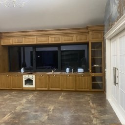 Respect Factory sp z o.o. - Kuchnia z zabudową meblową z jasnego drewna, wbudowany piekarnik, widoczne AGD, podłoga z płytek imitujących kamień, białe drzwi z przeszkleniami.