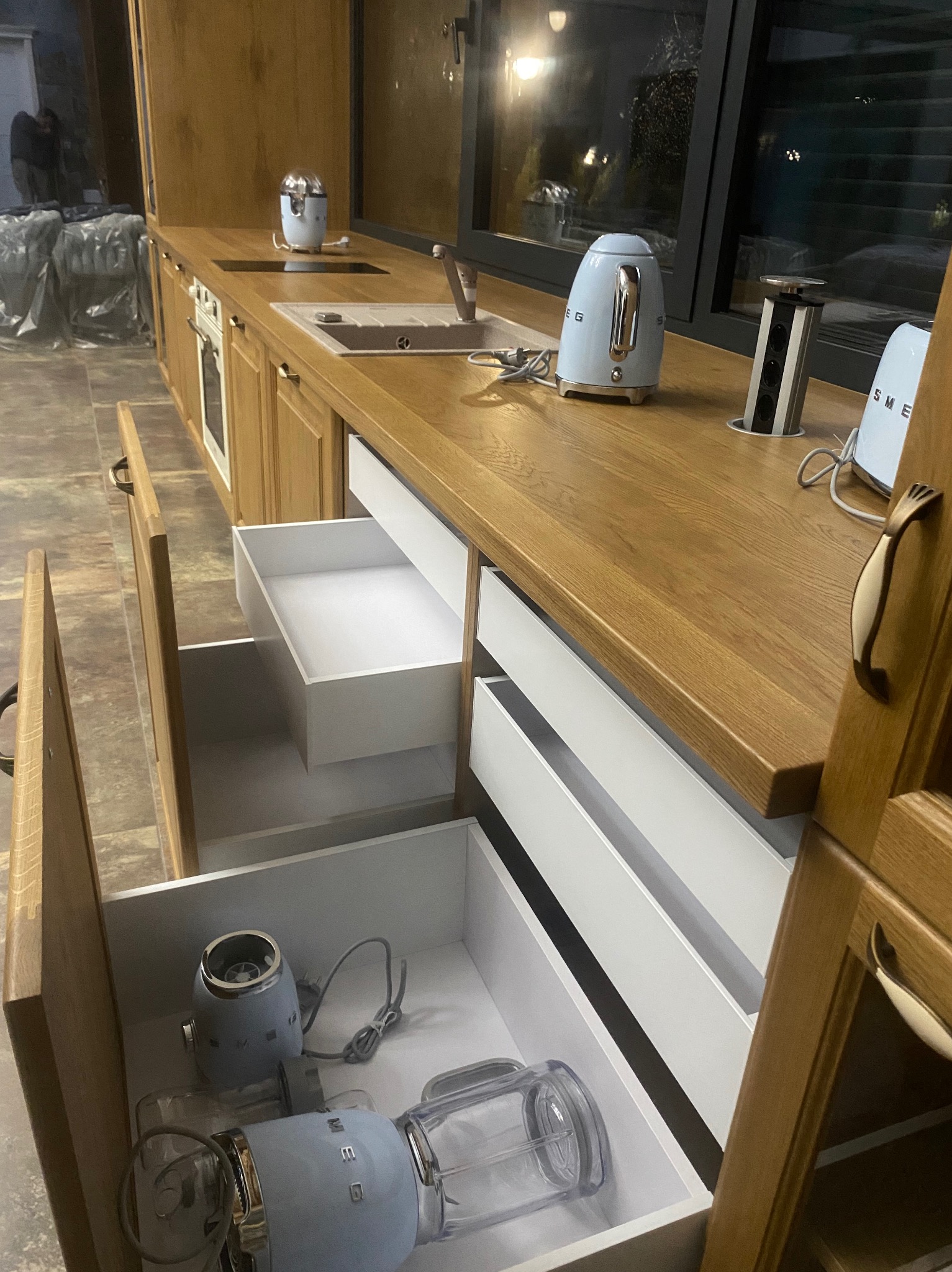 Nowoczesna kuchnia z drewnianymi frontami, białymi szufladami i sprzętem AGD marki Smeg w pastelowych odcieniach błękitu, niektóre szuflady otwarte ukazują przechowywane urządzenia.