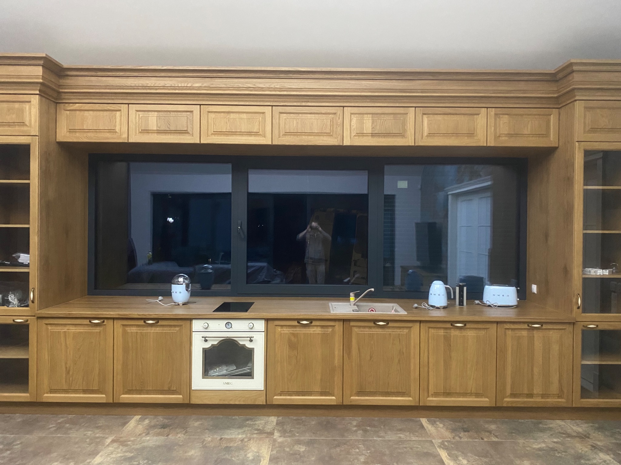 Klasyczna zabudowa kuchenna z drewnianymi szafkami, blatem i dużym oknem, z widocznym odbiciem osoby robiącej zdjęcie w szybie okna.