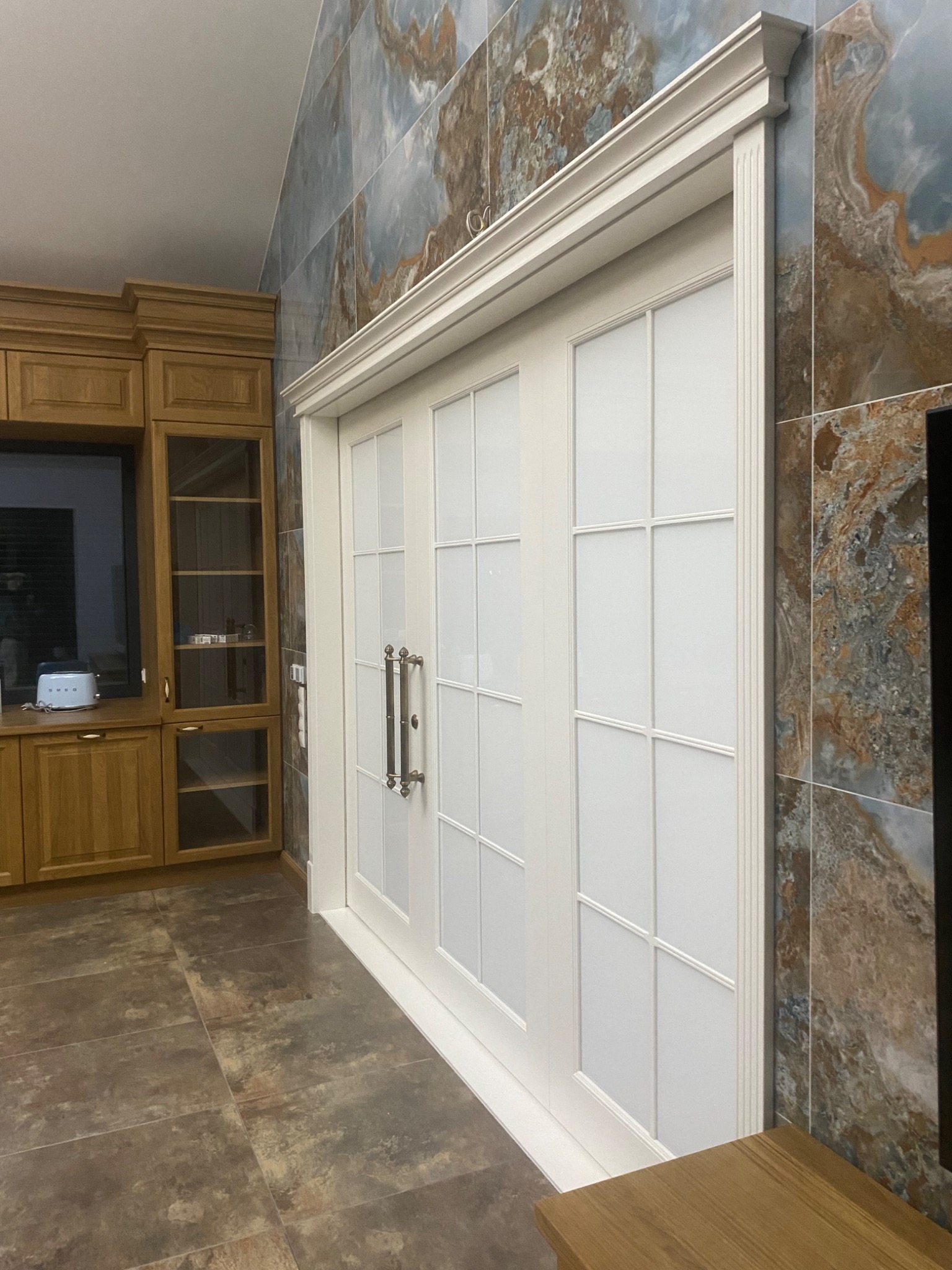 Białe, dwuskrzydłowe drzwi z dekoracyjnymi szprosami i ozdobną ramą, zamontowane w Kartuzach, widoczne na tle ściany wyłożonej płytkami imitującymi marmur, obok zabudowa kuchenna z drewna.