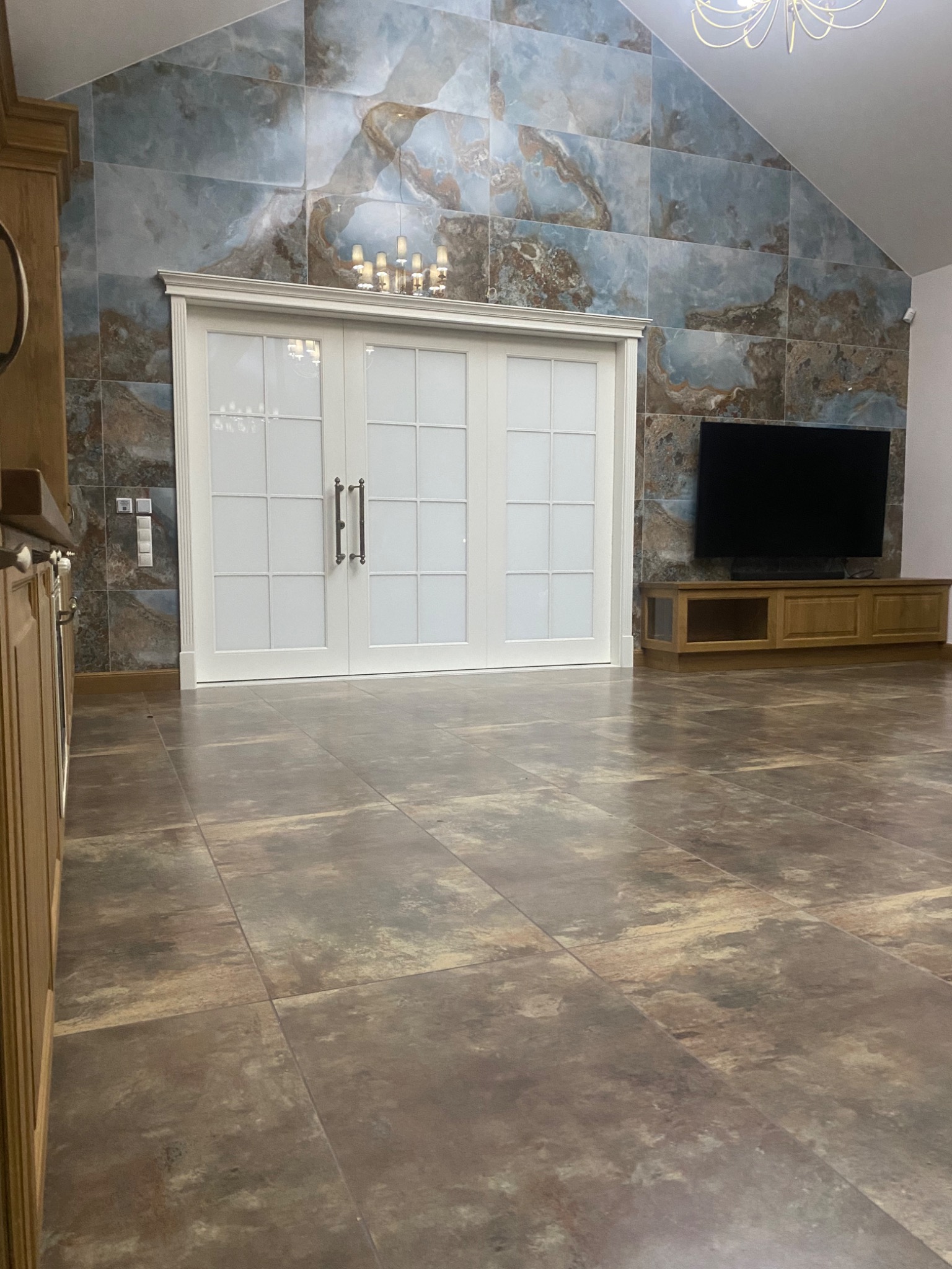 Nowoczesny salon z przesuwnymi drzwiami z białymi ramami, dekoracyjną ścianą z płytek imitujących marmur, drewnianą szafką RTV i podłogą z płytek w odcieniach brązu.