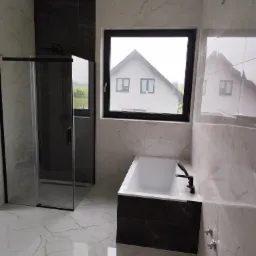 Nowoczesna łazienka z kabiną prysznicową, wanną i oknem z widokiem na dom. Białe marmurowe płytki z ciemnymi akcentami. Czarna armatura i rama okienna.