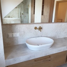 Ergo System Sp. z o.o. - Nowoczesna łazienka z owalną umywalką nablatową, złotą armaturą i lustrem odbijającym kabinę prysznicową oraz drewniane szafki. Wykończenie w jasnych, naturalnych barwach.