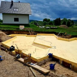 Fundamenty domu jednorodzinnego w trakcie budowy, ocieplone styropianem, z widocznymi narzędziami i elementami konstrukcyjnymi na tle domu i pochmurnego nieba.