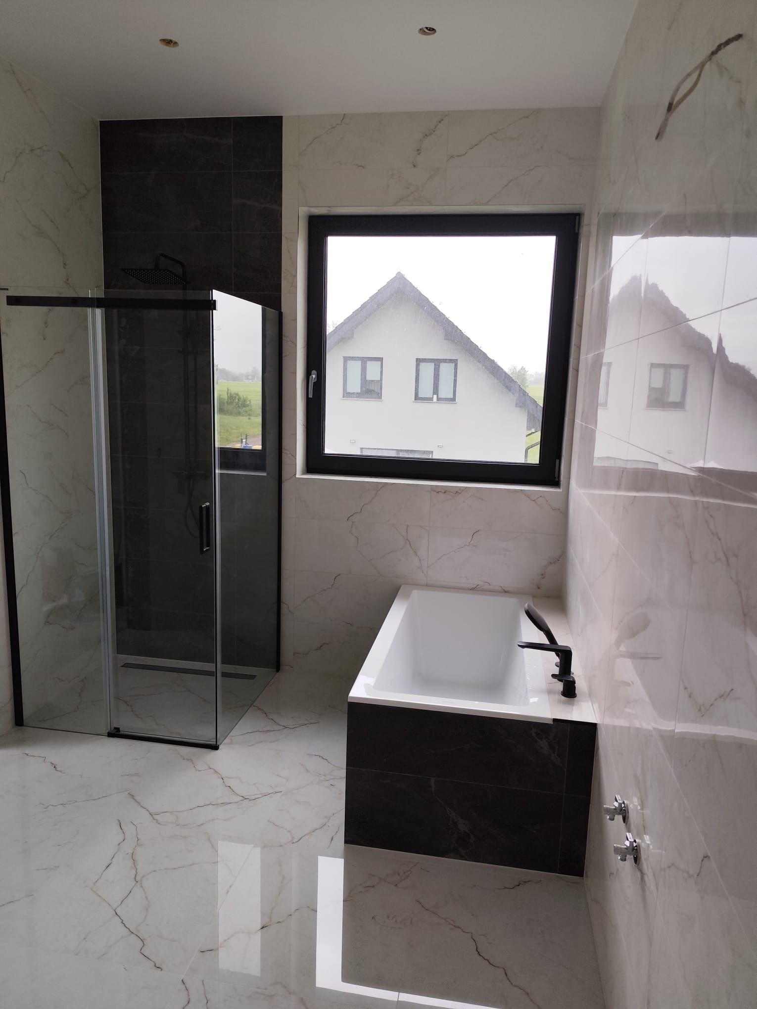 Nowoczesna łazienka z kabiną prysznicową, wanną i oknem z widokiem na dom. Białe marmurowe płytki z ciemnymi akcentami. Czarna armatura i rama okienna.