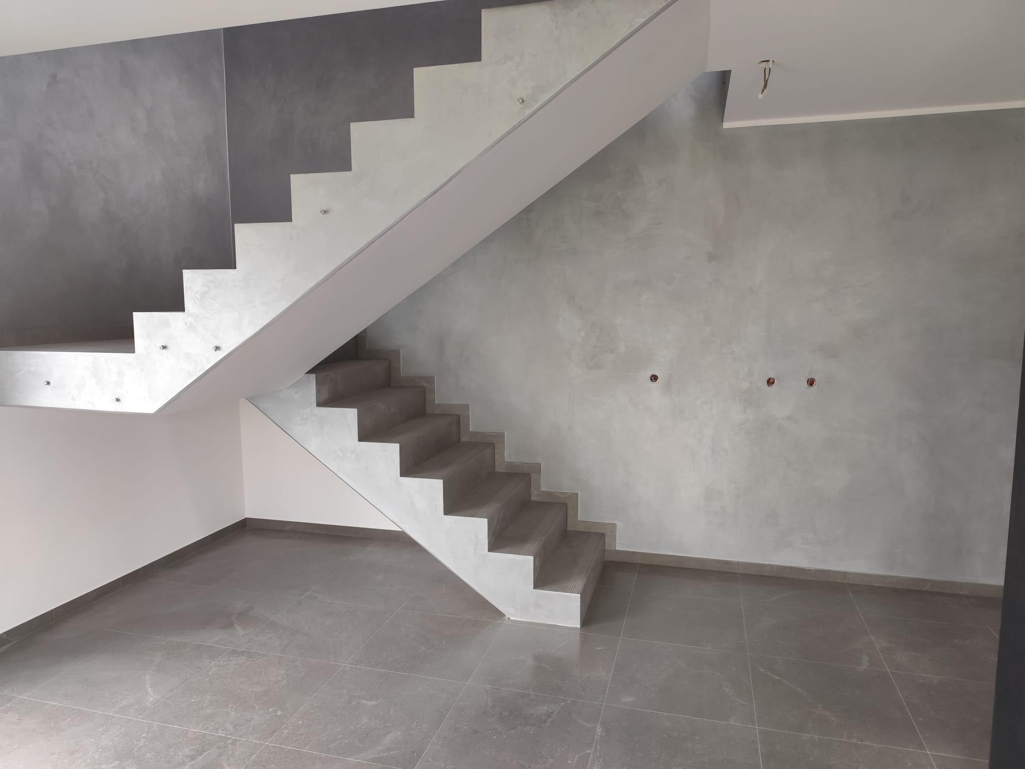 Wnętrze po remoncie: szare płytki podłogowe, ściany z efektem dekoracyjnym i nowoczesne schody z oświetleniem punktowym. Widoczne gniazdka elektryczne.