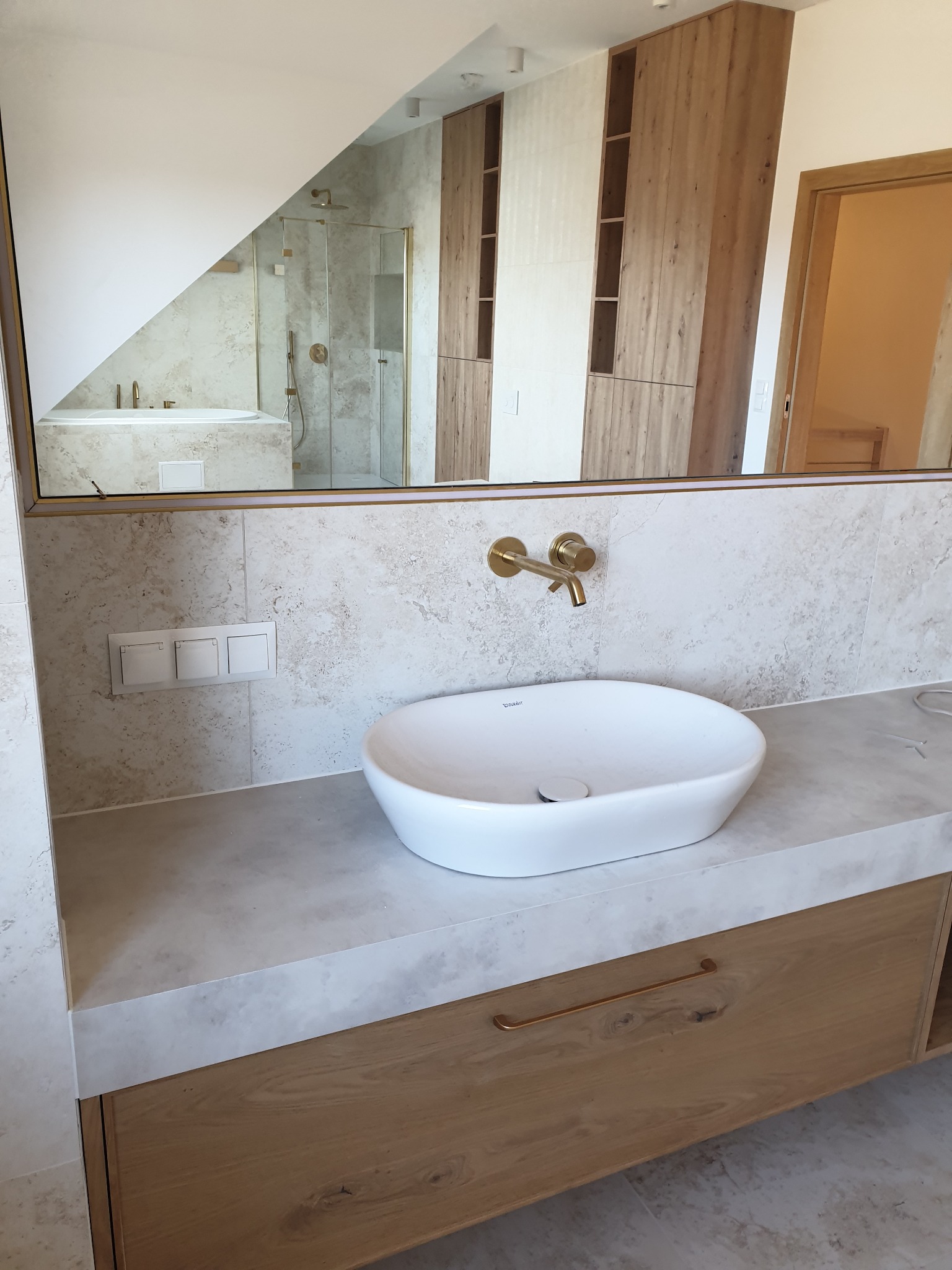 Nowoczesna łazienka z owalną umywalką nablatową, złotą armaturą i lustrem odbijającym kabinę prysznicową oraz drewniane szafki. Wykończenie w jasnych, naturalnych barwach.