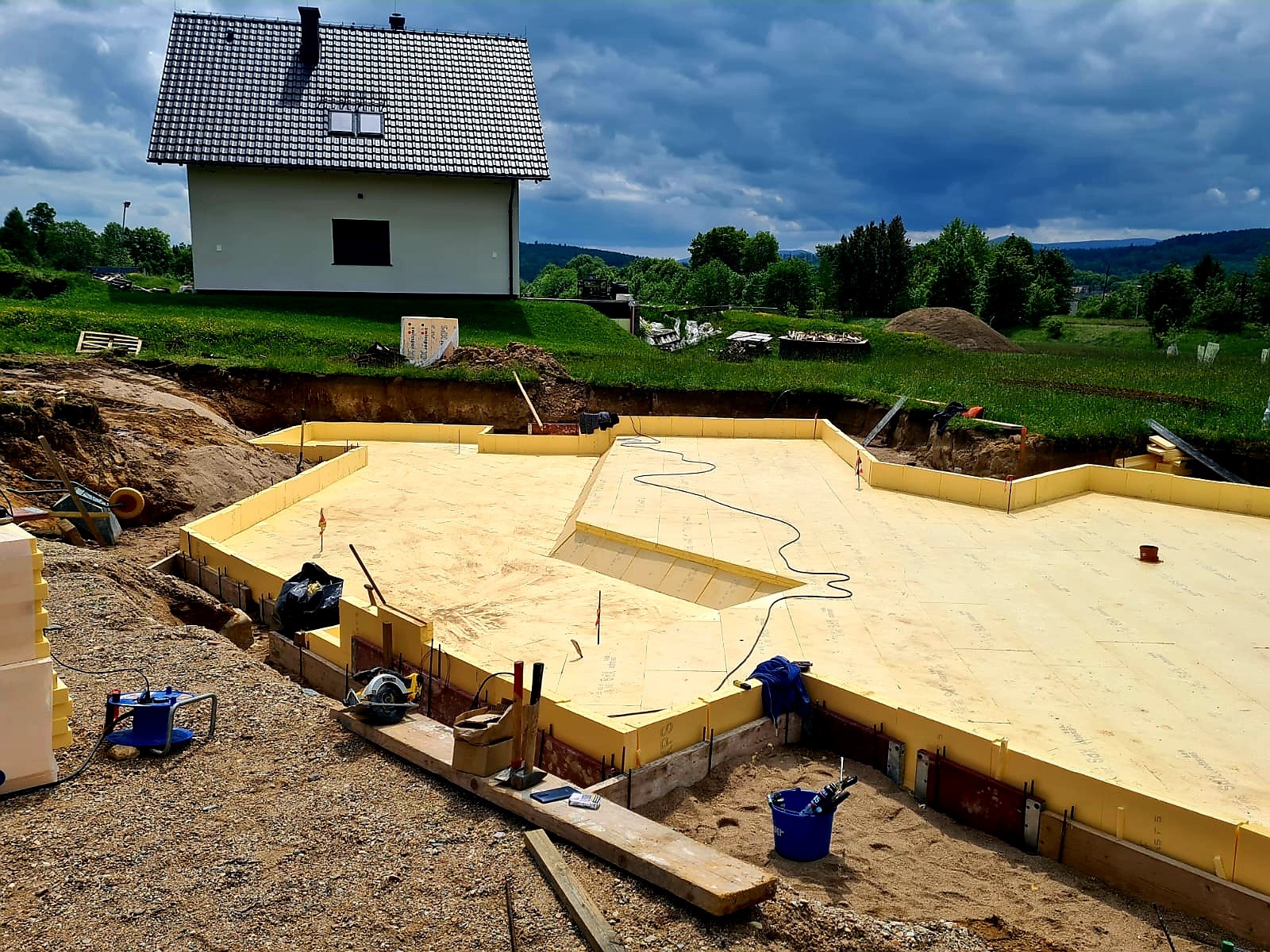 Fundamenty domu jednorodzinnego w trakcie budowy, ocieplone styropianem, z widocznymi narzędziami i elementami konstrukcyjnymi na tle domu i pochmurnego nieba.