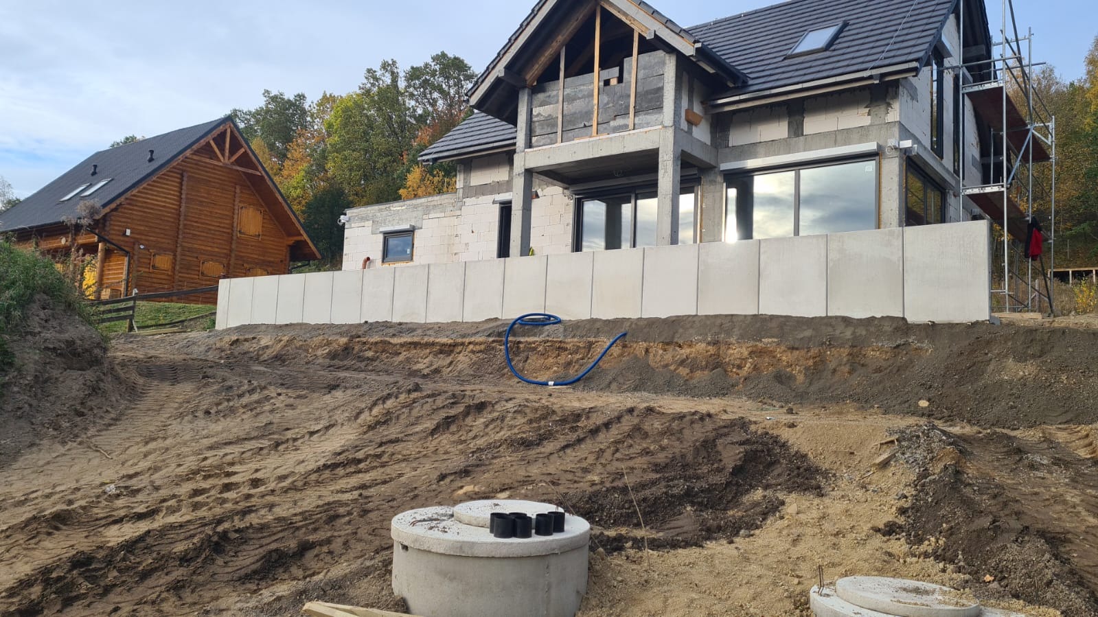 Budowa domu z betonu komórkowego, widoczna ściana oporowa z prefabrykatów. W tle dom z bali i rusztowania. Niebieski przewód na wykopie.
