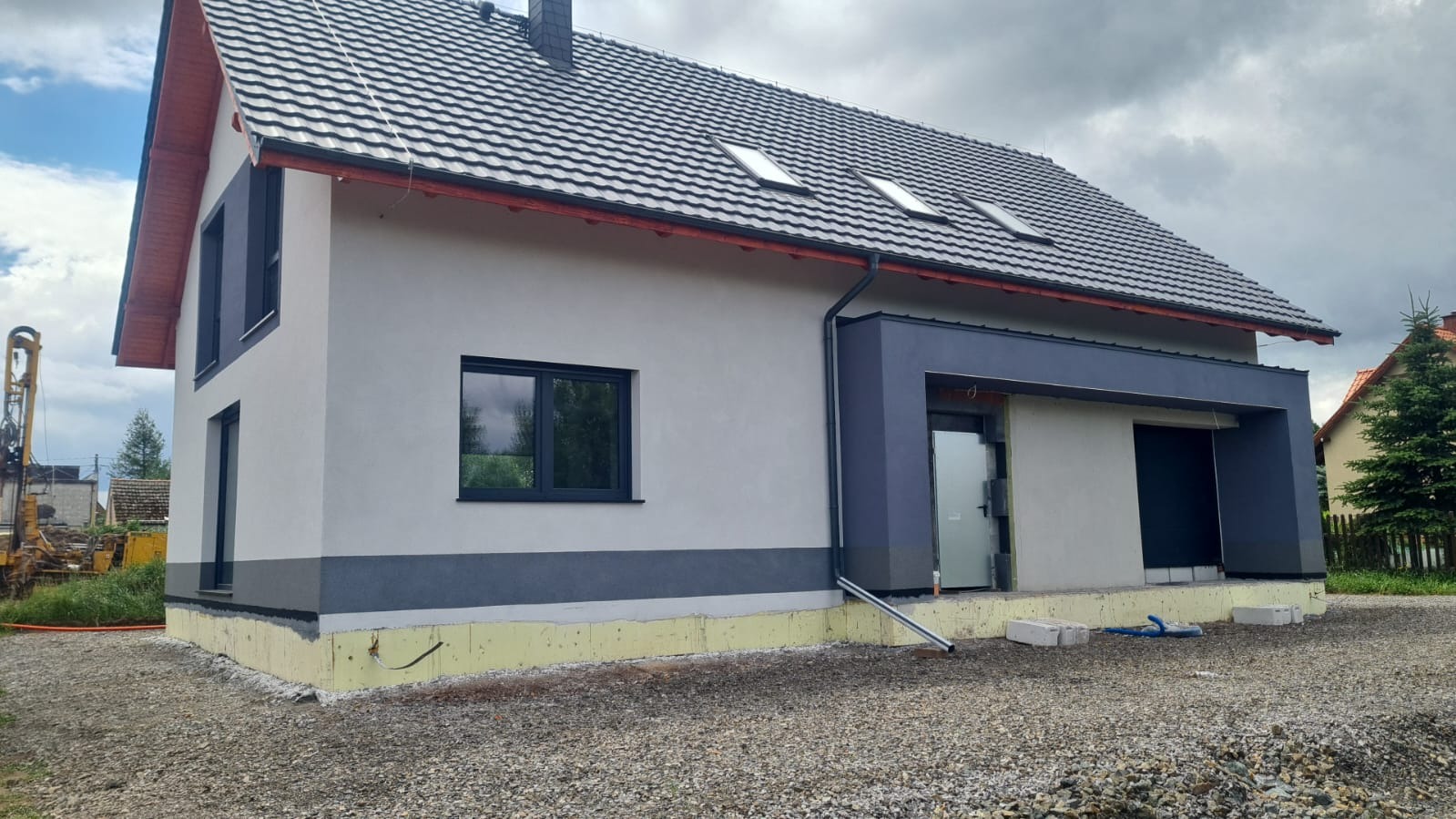 Nowoczesny, dwukondygnacyjny dom z ciemną stolarką okienną i grafitowym dachem. Widoczna elewacja w odcieniach szarości i wykończenie fundamentów. Na pierwszym planie żwirowe podłoże.