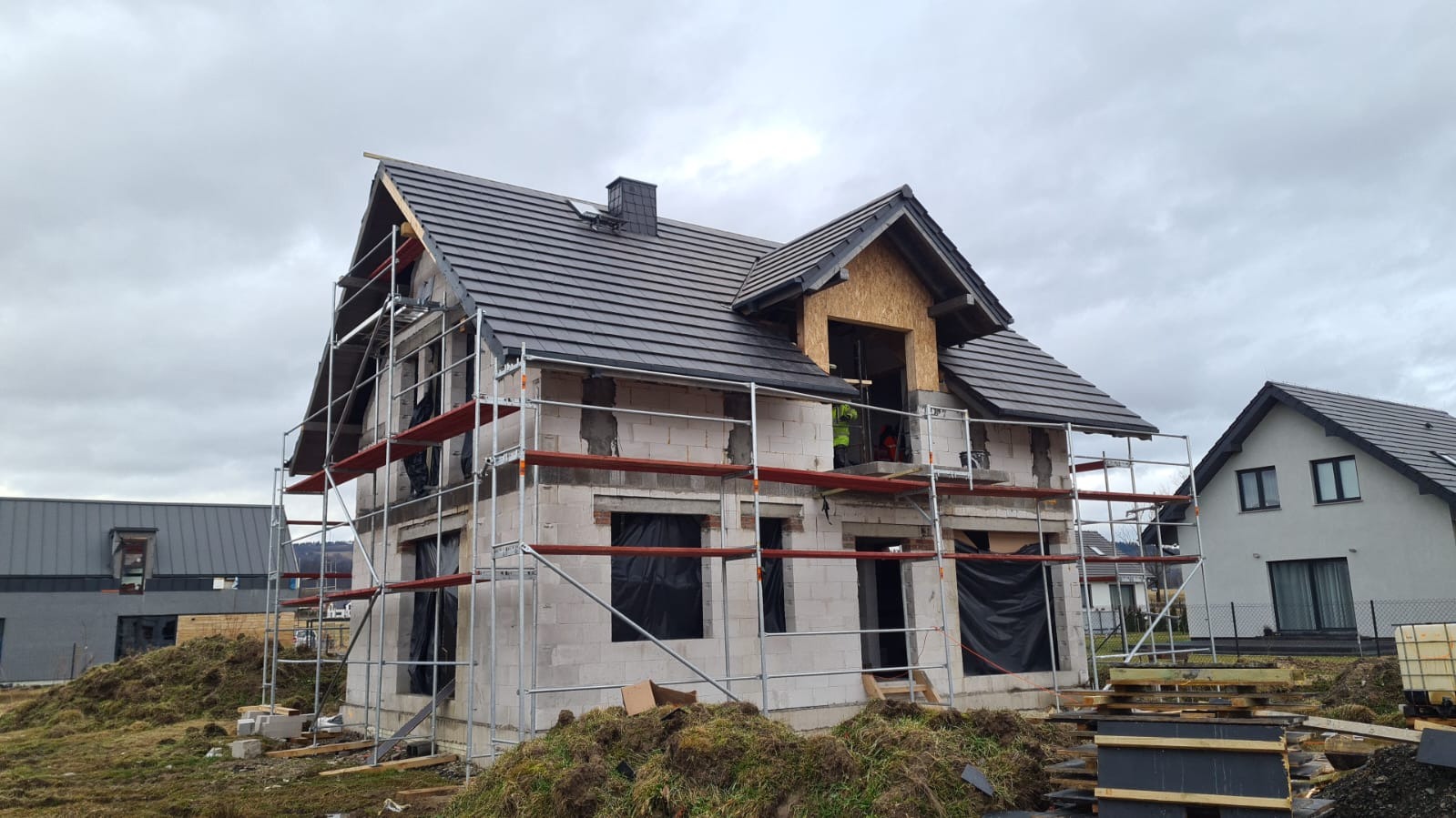 Niedokończony dom z białych bloczków, z ciemnym dachem, otoczony rusztowaniami. Widoczny komin i okno na poddaszu. W tle inne domy i pochmurne niebo.