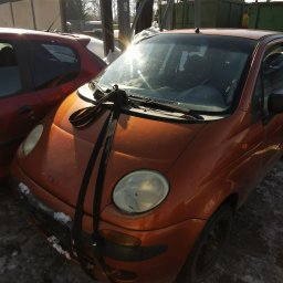 Metalic Spółka Jawna Janowiak - Pomarańczowy samochód z czarnymi pasami transportowymi na masce, stojący na placu z resztkami śniegu, w tle widać kontener i podnośnik samochodowy.