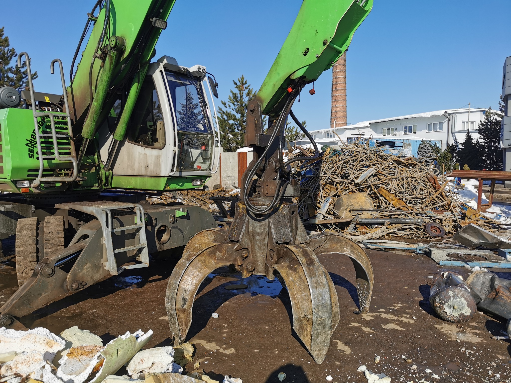 Zielona maszyna z chwytakiem hydraulicznym na tle sterty złomu metalowego i ceglanego komina