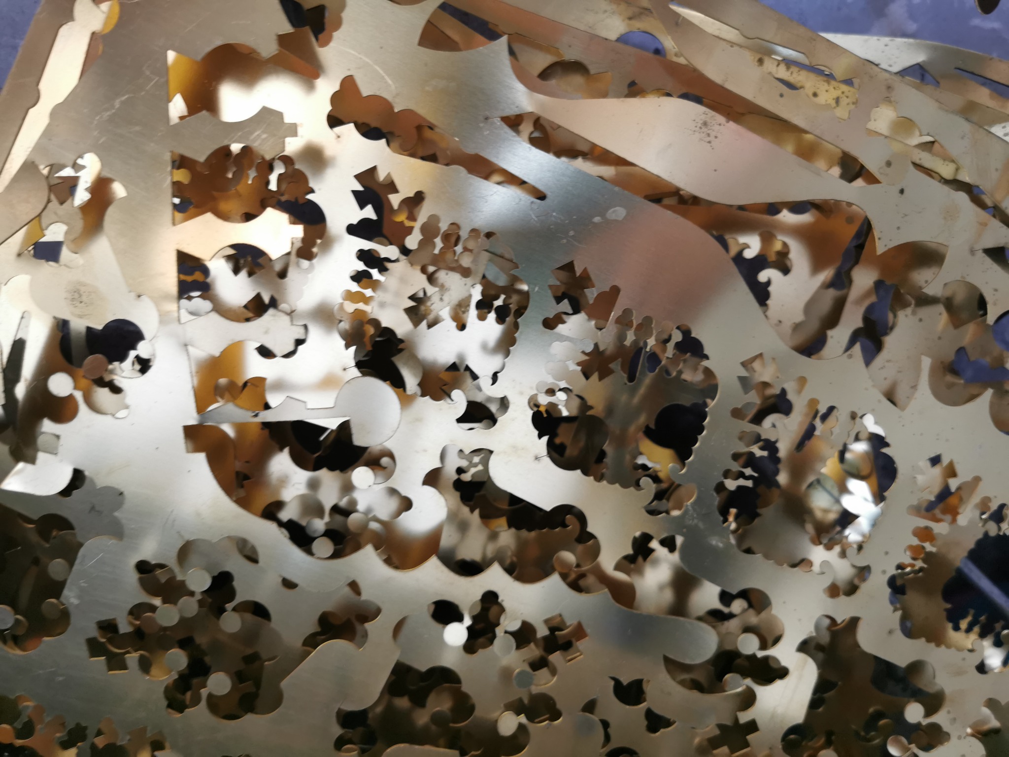 Metalowy złom z wyciętymi wzorami, widoczne nieregularne kształty i otwory, oświetlenie tworzy refleksy na powierzchni.