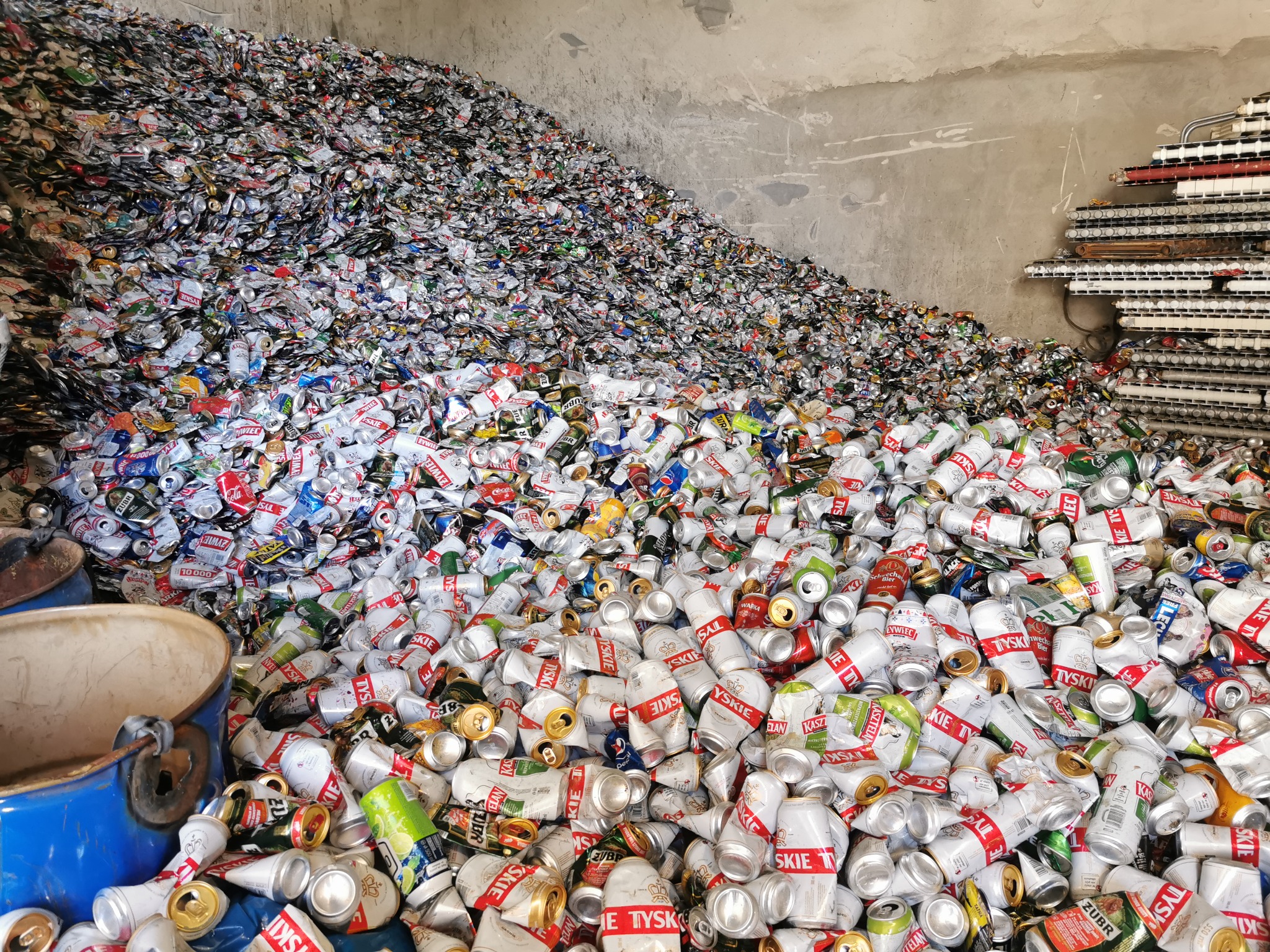 Góra zgniecionych, aluminiowych puszek po napojach, głównie piwa, w magazynie z betonowymi ścianami. Widoczny niebieski beczkowóz i elementy instalacji na ścianie.
