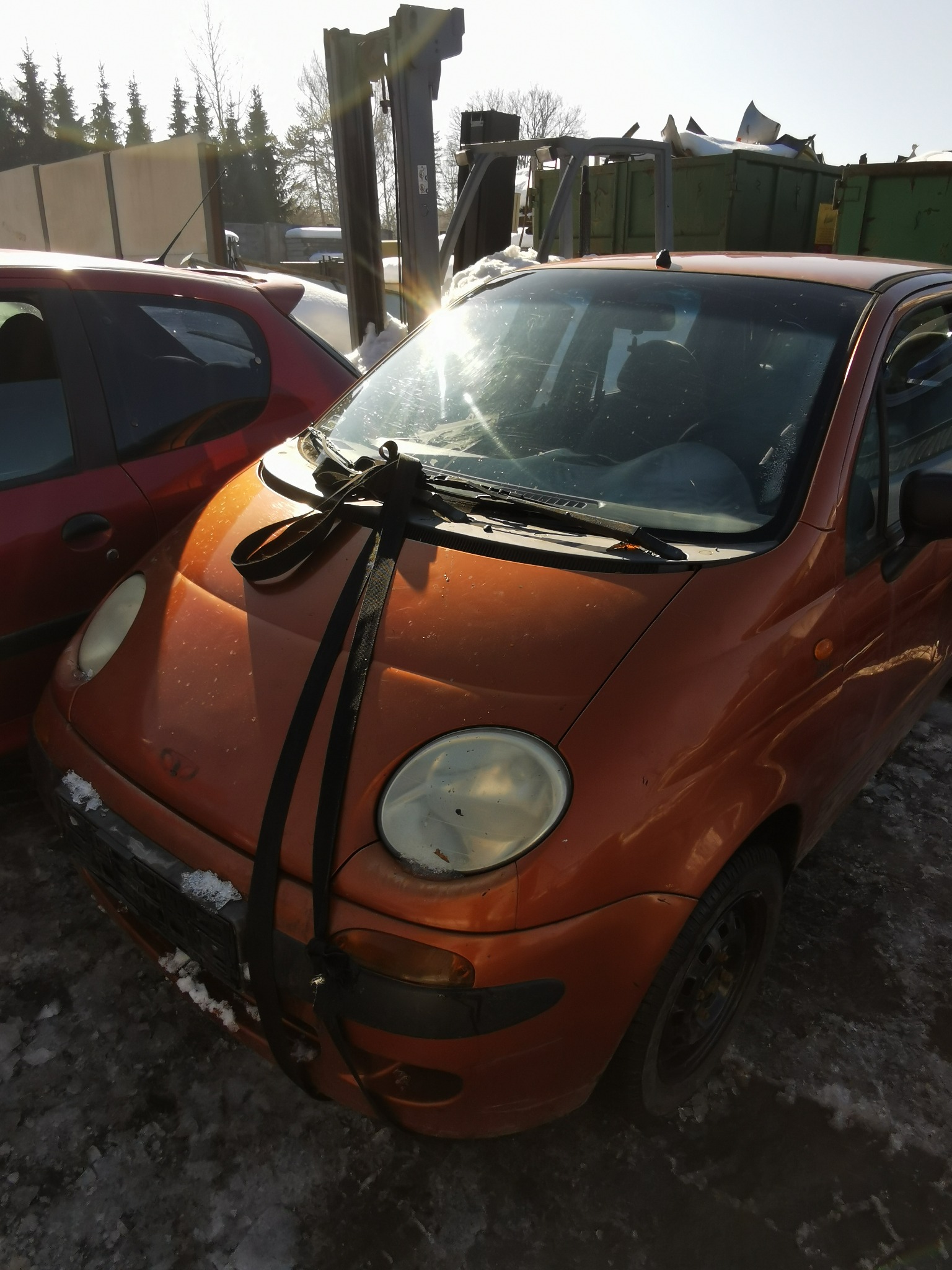 Pomarańczowy samochód z czarnymi pasami transportowymi na masce, stojący na placu z resztkami śniegu, w tle widać kontener i podnośnik samochodowy.