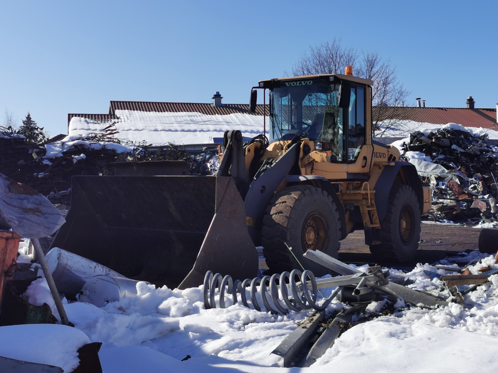 Żółta ładowarka Volvo na placu złomowym, z kupą metalowych odpadów i śniegiem w tle, widoczne sprężyny zawieszenia i fragmenty karoserii.