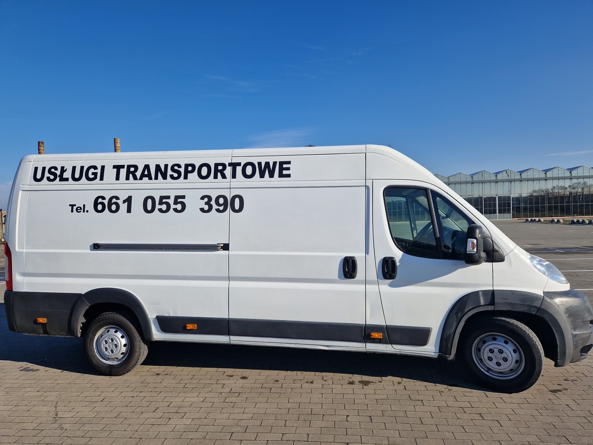Biały samochód dostawczy z czarnym napisem 'USŁUGI TRANSPORTOWE' i numerem telefonu, zaparkowany na brukowanym placu, z widoczną szklarnią w tle.