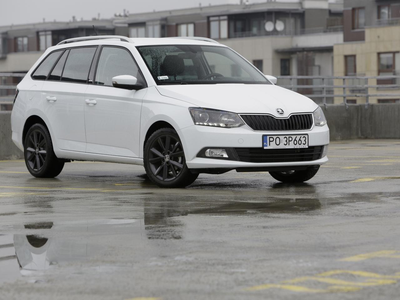 Biała Skoda Fabia Combi z czarnymi felgami na mokrym parkingu wielopoziomowym, widok z przodu pod kątem.