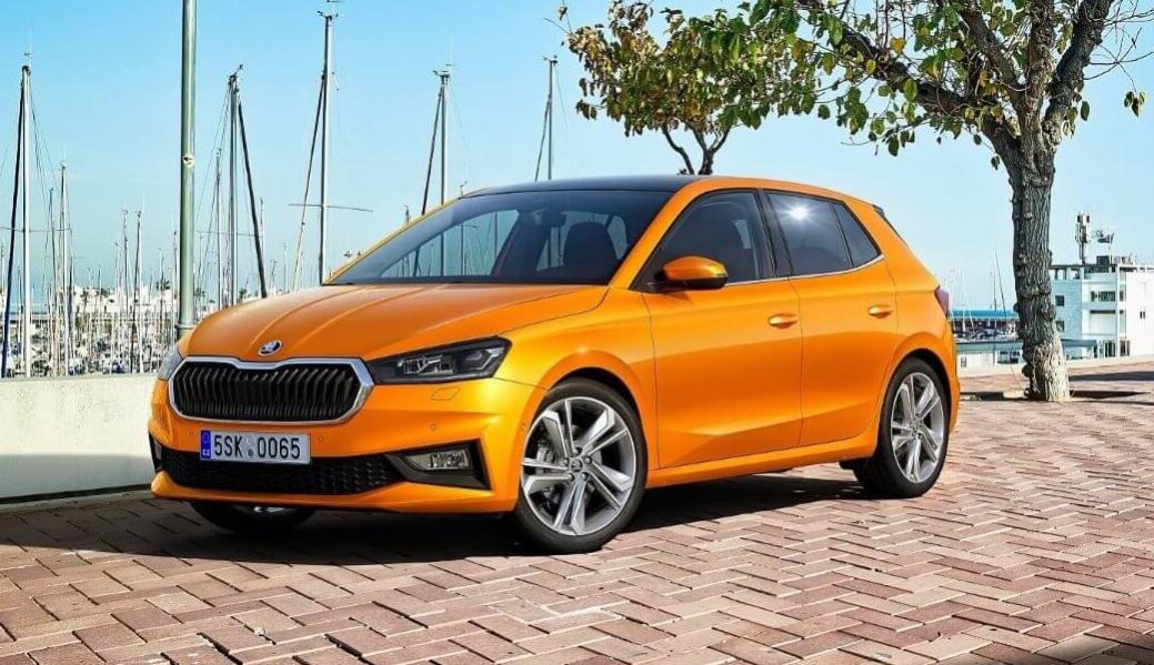 Pomarańczowa Skoda Fabia hatchback zaparkowana na brukowanym placu z widokiem na port jachtowy w tle, z drzewem po prawej stronie kadru.