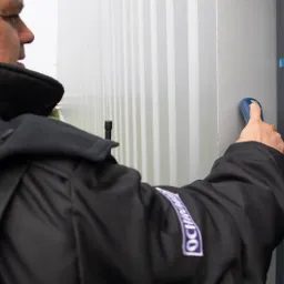 Ochroniarz w czarnej kurtce z logo firmy, używający niebieskiego czytnika zbliżeniowego na tle szarej, pionowo żebrowanej ściany kontenera.