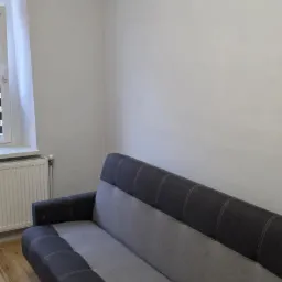 Jasnoszary pokój po malowaniu z szarą sofą. Widoczne gładkie ściany, okno i drewniana podłoga. Minimalistyczny wystrój po remoncie.