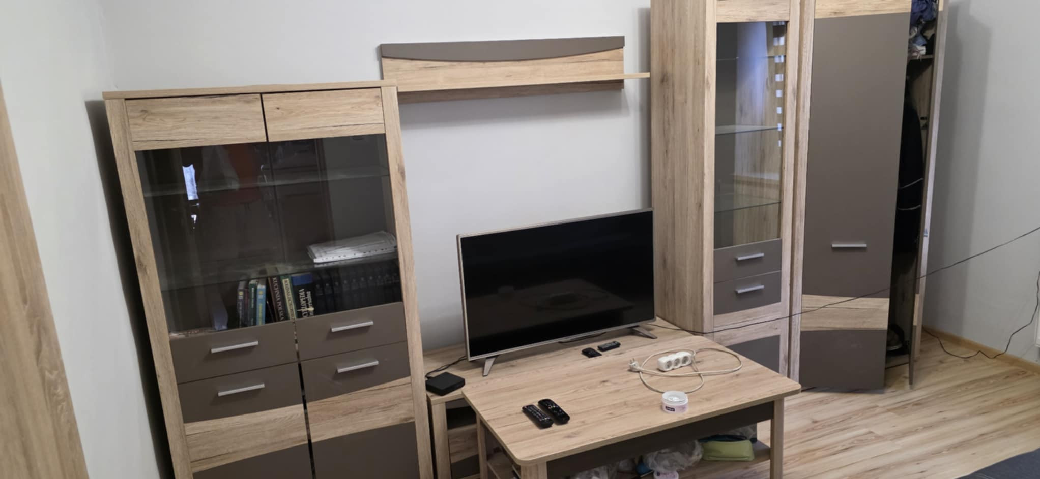 Ukończony pokój po remoncie: meble w odcieniach drewna i szarości, telewizor na stoliku. Ściany pomalowane na biało. Widoczne ślady po remoncie na podłodze.